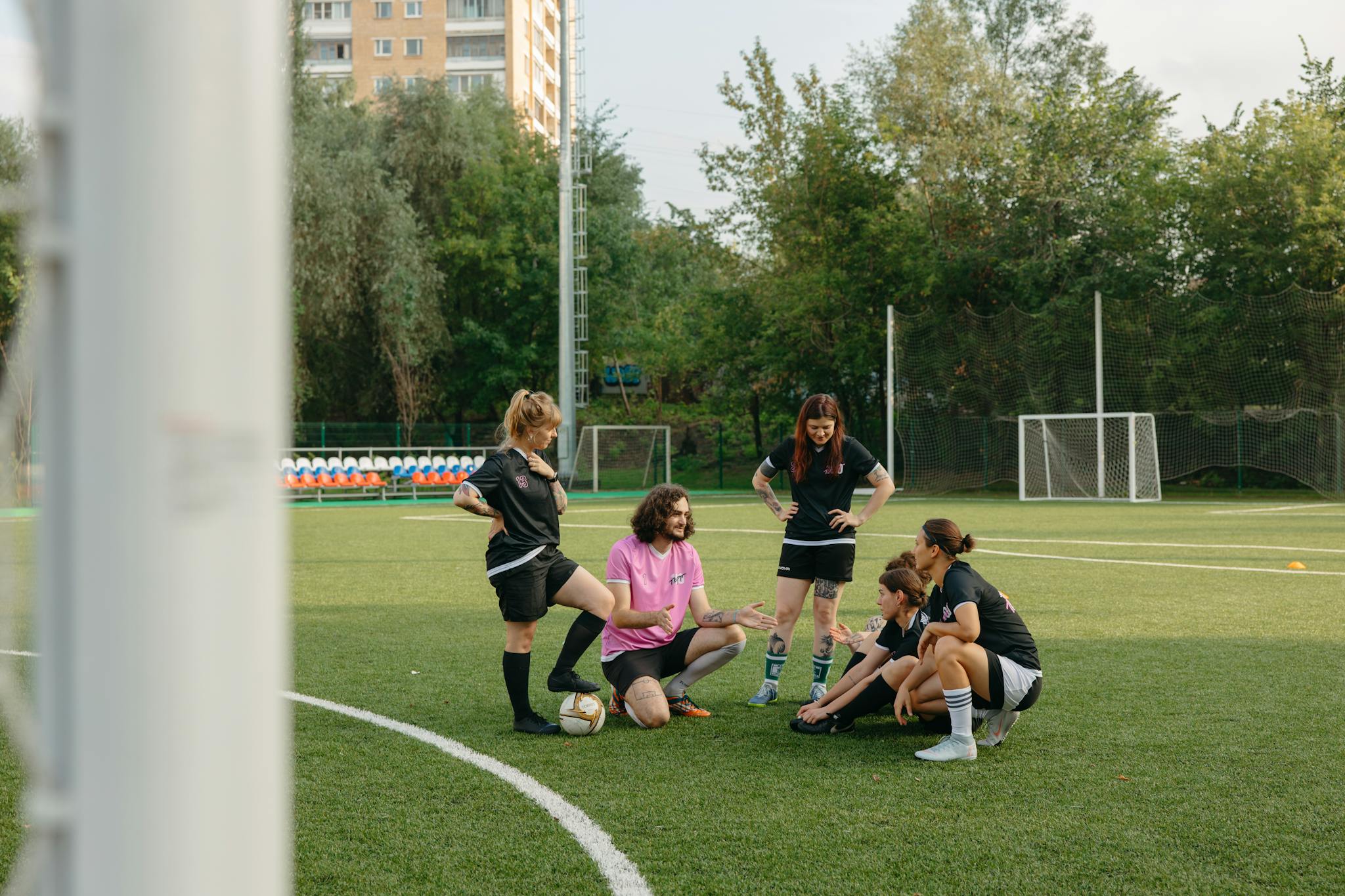 A mixed group of adults gathers on a sunny day for a team discussion on an outdoor football field.